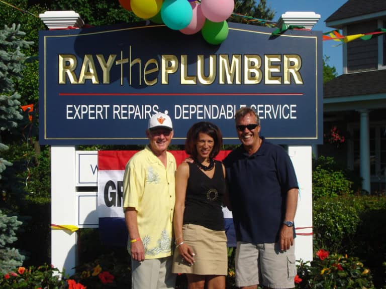 Three adults stand smiling in front of a “Ray the Plumber” business sign outdoors, decorated with balloons and a banner, on a sunny day, creating the perfect scene for an about us introduction.