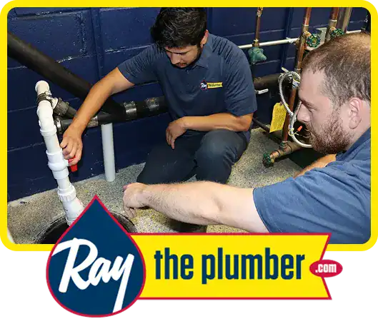 Two plumbers in blue uniforms work on a pipe system indoors, inspecting a valve. “Ray the plumber .com” logo is displayed at the bottom of the image.